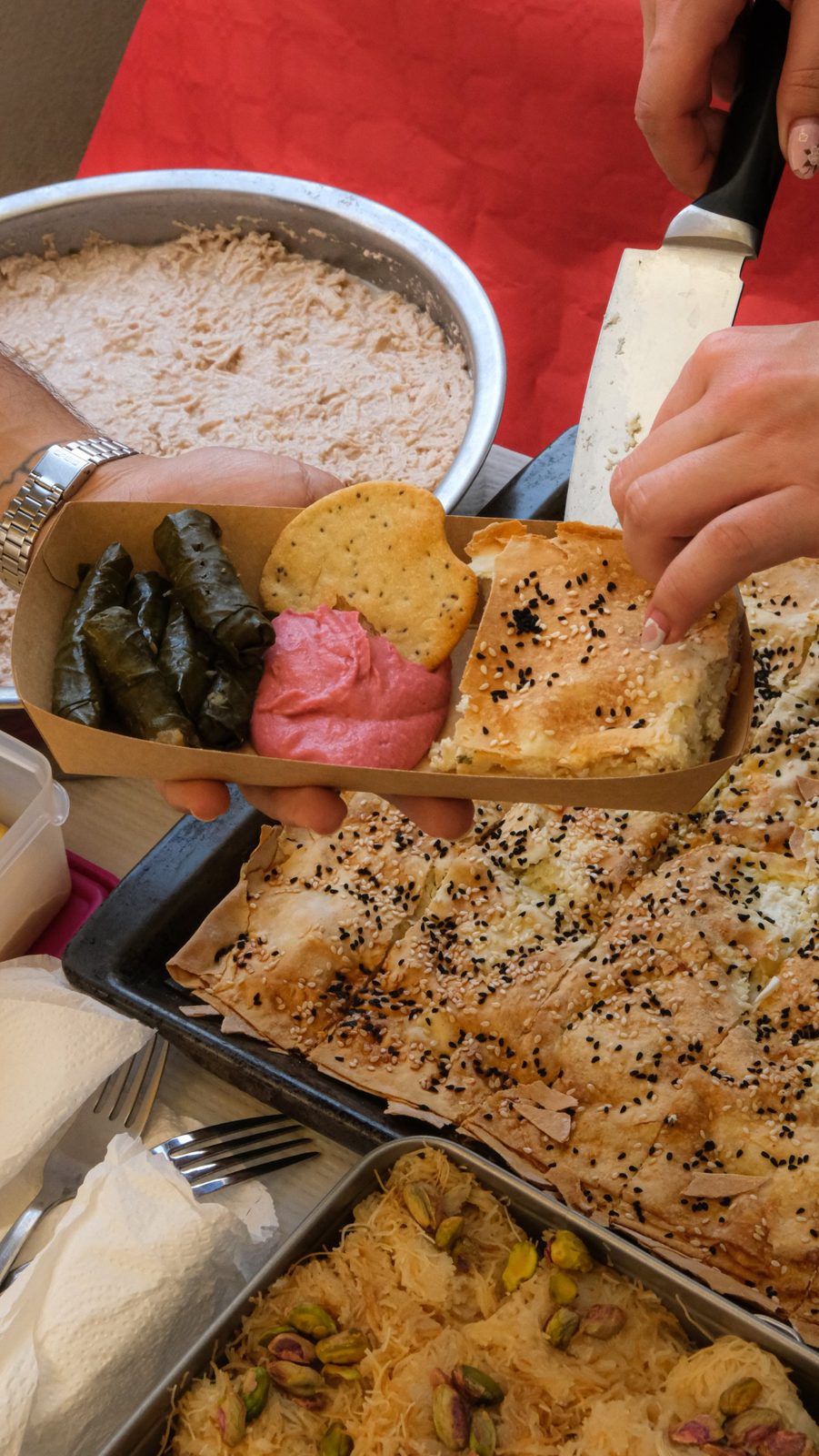 A takeaway box of sarma, pink hummus, a cracker and börek, with trays of börek and kadayıf behind